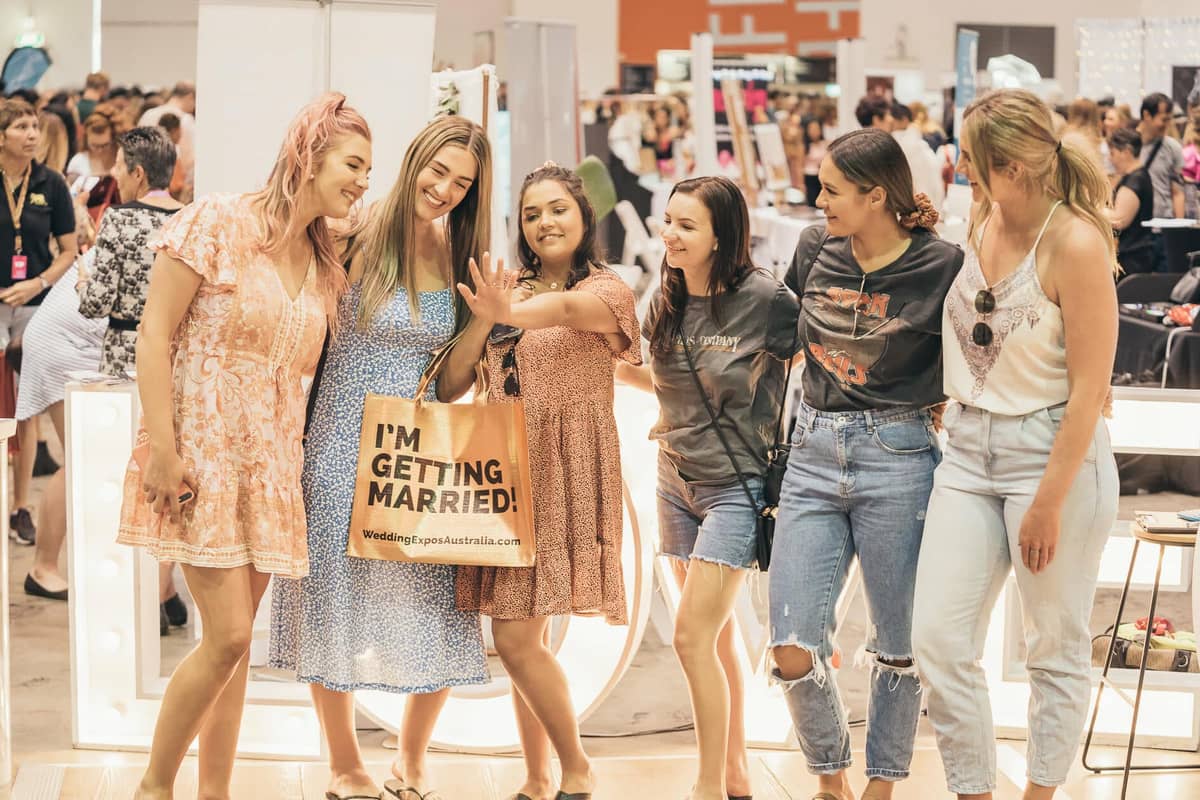 Six women gathered around a Gold “I'm Getting Married” Bag at a wedding expo